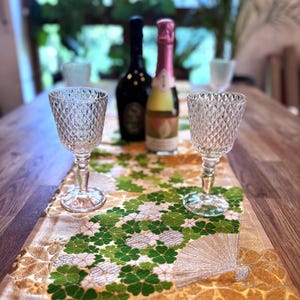 Könnte beinhalten: Eine Tischdekoration mit zwei Weingläsern mit Diamantmuster, einem dekorativen Tischläufer mit grünen Blumen- und Fächermotiven und zwei Weinflaschen. Der Läufer ist ca. 152 cm lang.