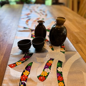 Könnte beinhalten: Ein Tischläufer mit floralem Design, mit hellblauem Hintergrund und bunten gestickten Blumen. Zwei kleine schwarze Schalen und zwei dunkelbraune Keramikflaschen befinden sich auf dem Läufer. Der Tisch ist aus hellem Holz.