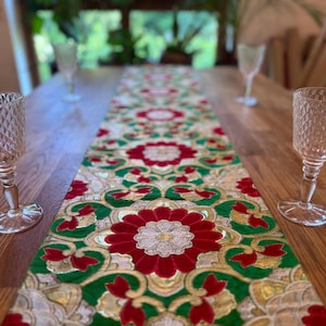 Könnte beinhalten: Ein Holztisch ist mit einem dekorativen Tischläufer und zwei Kristallkelchen gedeckt. Der Läufer zeigt ein sich wiederholendes Blumenmuster in Rot, Grün und Gold. Die Kelche haben ein Rautenmuster.