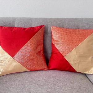Könnte beinhalten: Zwei quadratische Dekokissen mit geometrischem Color-Blocking-Design. Jedes Kissen kombiniert einfarbigen roten Stoff, ein strukturiertes orangefarbenes Paneel mit wiederkehrendem Webmuster und einen glatten goldfarbenen dreieckigen Bereich.