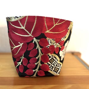 Könnte beinhalten: Ein kleiner Stoffbehälter mit floralem Design. Der Behälter hat einen schwarzen Hintergrund mit roten Blättern und Beeren, akzentuiert mit goldenen und weißen Details. Er steht auf einer Holzoberfläche.
