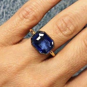 May include: A gold ring featuring a large, rectangular, faceted deep blue gemstone set in a simple metal band. The stone has a clear, polished surface with sharp edges and a vibrant, saturated blue color, reflecting light across its facets.