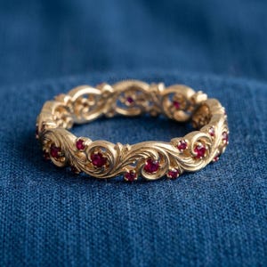 Puede incluir: Un anillo de banda de oro con intrincados patrones de volutas y rubíes rojos redondos incrustados. La superficie metálica tiene un acabado texturizado y el diseño incluye motivos de remolinos repetidos que rodean toda la circunferencia de la joya.