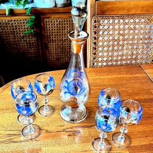 May include: A vintage glass decanter with a crystal stopper and six matching cordial glasses. The clear glass is decorated with blue floral designs and gold trim. The set is displayed on a wooden table.