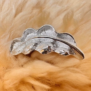 Peut inclure: Une broche argentée en forme de feuille. La broche présente une surface texturée et détaillée, sur un fond brun clair et doux. Un accessoire décoratif.