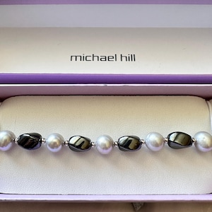 May include: A bracelet featuring alternating white spherical pearls and dark gray metallic-toned beads, separated by small silver-colored spacers. The jewelry is presented inside a light purple box with the text michael hill printed in black.
