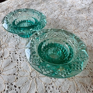 May include: Two teal-colored glass candle holders with diamond-cut patterns. The candle holders have a circular shape with a central well for holding candles. The glass has a slight green tint, and the edges are rounded.