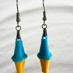 May include: A pair of earrings featuring a blue and yellow cone design. The earrings have a silver-colored hook and a spring detail. The cone shape has a blue top and a yellow bottom. The earrings are displayed against a white background.
