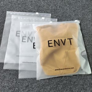 Op de afbeelding: Drie doorschijnende, matglazen plastic zakken met ritssluiting op elkaar gestapeld. Elke zak is voorzien van de zwarte tekst "ENVT" in een vet schreefloos lettertype, samen met de website "www.EnavantActive.com" onderaan. Een geel kledingstuk is zichtbaar.