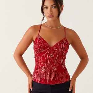 Op de afbeelding: Een rode camisole top met een V-hals en spaghettibandjes. De top is versierd met bloemenborduursels en kralen in rood, goud en wit. De onderste helft van de top heeft verticale kralenfranjes. Het model draagt een zwarte broek.