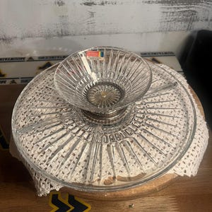 May include: A clear glass serving dish featuring a central raised bowl attached to a wider, flat circular base with a divided section design. The glass has a radial ribbed pattern and includes a small rectangular label reading Heritage.