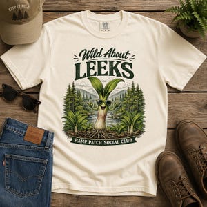 Puede incluir: Camiseta color crema con la frase "Wild About Leeks" y un personaje de puerro de dibujos animados con gafas de sol. El gráfico incluye una escena de montaña y las palabras "Ramp Patch Social Club". También se ven una gorra de béisbol, gafas de sol y vaqueros.