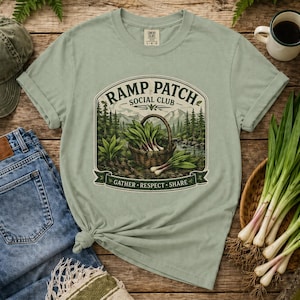 May include: Sage green t-shirt featuring a graphic of a basket of ramps with the text "RAMP PATCH SOCIAL CLUB". The design includes a forest scene and the words "GATHER, RESPECT, SHARE". The shirt is styled with a denim jeans, a green cap, and a mug.