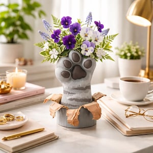 Puede incluir: Un jarrón gris con forma de pata de gato que contiene un ramo de flores moradas, blancas y verdes. El jarrón tiene un efecto de papel rasgado alrededor de la base. También se ven una taza de café, una vela encendida y anillos dorados.