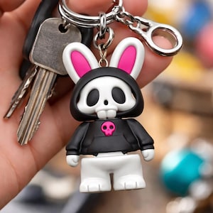 Könnte beinhalten: Ein neuartiger Schlüsselanhänger mit einem weißen Hasenschädel-Charakter mit rosa Innenohren, der einen schwarzen Kapuzenpullover mit einem rosa Totenkopf-Emblem trägt. Der Schlüsselanhänger ist an einem silbernen Schlüsselring mit Schlüsseln befestigt.