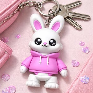 Könnte beinhalten: Ein weißer Hasen-Schlüsselanhänger mit rosa Akzenten, der einen rosa Kapuzenpullover trägt. Der Schlüsselanhänger ist an einem silbernen Schlüsselring mit Schlüsseln befestigt. Der Hintergrund ist eine hellrosa Oberfläche mit herzförmigem Glitzer.