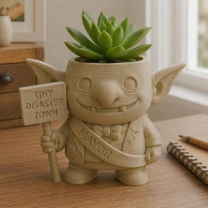 May include: A beige, goblin-like planter featuring a succulent, wearing a sash labeled MAYOR, and holding a sign that reads TINY DISASTER TOWN. The figure has large ears, a wide grin with visible teeth, and is dressed in a suit with a bow tie.