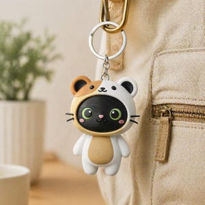 May include: A novelty cat keyring with a black face and green eyes, wearing a white and brown bear costume. The keyring is attached to a silver ring and a gold-coloured clasp. The cat has a small pink nose and whiskers.