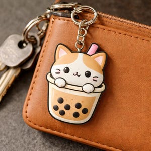 Op de afbeelding: Een schattige sleutelhanger met kattenmotief, met een cartoonkat in een boba-theekopje. De sleutelhanger is bevestigd aan een zilveren sleutelring en staat tegen een bruine leren portemonnee. De kat heeft oranje en witte vacht en de boba-thee heeft zwarte tapiocaparels.