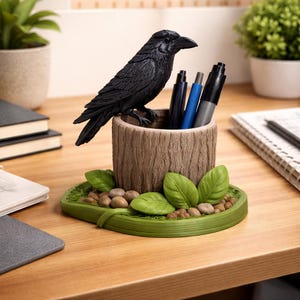 Puede incluir: Un portalápices de cuervo negro con diseño de tronco de árbol. El portalápices tiene una base verde con hojas y pequeñas piedras. Varios bolígrafos y rotuladores están dentro. El portalápices está sobre un escritorio de madera.