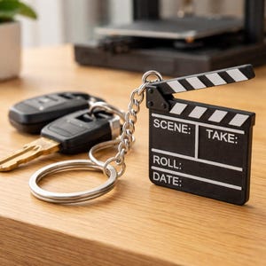 May include: A black and white film clapperboard keychain with silver chain and key rings. The clapperboard has the words "SCENE:" "TAKE:" "ROLL:" and "DATE:" printed on it. Car keys are attached to the key rings. The keychain is on a wooden surface.