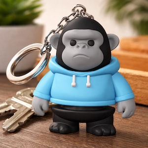 May include: A keychain featuring a stylized gorilla figure with a black head, gray face and ears, and a light blue hoodie. The keychain has a silver chain and ring, and the gorilla is wearing black pants and shoes.