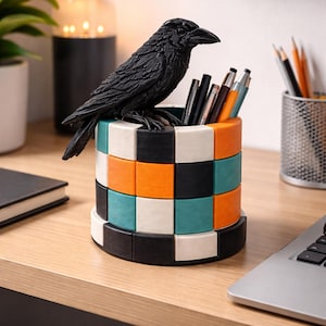 May include: A decorative desk organizer featuring a black raven perched atop a cylindrical container. The container is composed of stacked, colorful squares in black, white, orange, and teal. Pens and pencils are visible inside the organizer.