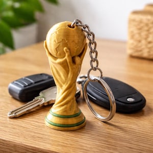 May include: A gold-coloured keychain with a trophy design, depicting a globe held by hands. The keychain is attached to a silver chain and ring. Other keys and a car key fob are on a wooden surface.