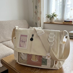Può includere: Una borsa a tracolla in tela color crema con una tasca frontale trasparente, uno scomparto verticale con cerniera e una piccola finestra rettangolare che mostra un'illustrazione di un orso. La borsa include una tracolla larga regolabile e un ciondolo a forma di coniglio.