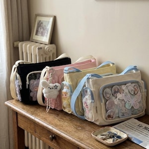 May include: A collection of colorful shoulder bags in black, pink, yellow, and cream, each with a clear pocket displaying various charms and accessories. The bags are arranged on a wooden table, with a small tray of keys and a newspaper in the foreground.
