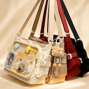 Può includere: Un set di tre borse a tracolla in tela nei colori crema, rosso e nero. La borsa color crema presenta una tasca frontale trasparente con adesivi e cartoline, decorata con stelle di peluche e un portachiavi a forma di orsetto peloso.