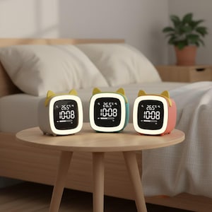 Könnte beinhalten: Drei digitale Wecker mit Katzenohren-Design in Grau, Türkis und Rosa stehen auf einem kleinen Holztisch. Jede Uhr zeigt die Uhrzeit 10:08 Uhr und die Temperatur 26,5 Grad Celsius an. Die Uhren haben einen weißen Rand und gelbe Katzenohren.