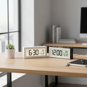 以下が含まれることがあります： 木製の机の上に2つのデジタル目覚まし時計があります。1つは白で「6:30」と表示され、もう1つは黒で「12:00」と表示されます。両方の時計には日付と時刻も表示されます。小さな鉢植えの植物とノートも机の上にあります。