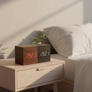 Peut inclure: Une horloge numérique en bois avec deux blocs carrés, un marron et un noir. Le bloc marron affiche l'heure en rouge, le bloc noir en blanc. Une petite plante est posée sur l'horloge, qui est sur une table de chevet. L'heure est 19h23 et 19h21.