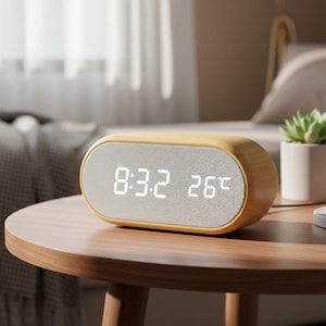 Könnte beinhalten: Ein digitaler Wecker aus Holz mit hellgrauer Lautsprecherabdeckung zeigt die Uhrzeit 8:32 und die Temperatur 26°C an. Die Uhr hat eine abgerundete rechteckige Form und steht auf einem Holztisch.