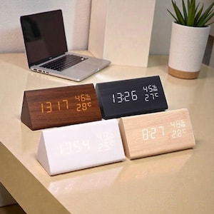 May include: Four wooden digital alarm clocks in various colors: dark brown, black, white, and light wood. Each clock displays the time, humidity, and temperature. A laptop and a potted plant are in the background.