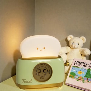 Puede incluir: Un despertador verde menta y dorado con forma de tostadora, con una luz en forma de tostada sonriente en la parte superior. El reloj muestra la hora 12:54 y la temperatura 20°C. La palabra "Toast" está impresa en la parte delantera.