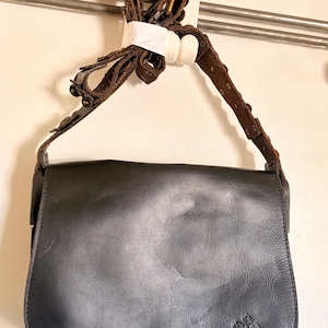 May include: Black leather crossbody bag with a flap closure and a brown, adjustable strap. The bag features a decorative floral emblem on the lower right corner and a brown leather strap with metal accents.
