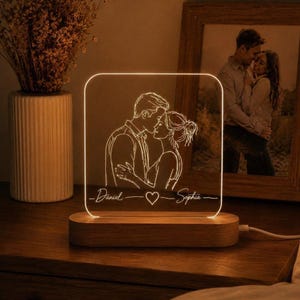 May include: A personalised night light with a couple's embrace design, the names "Daniel" and "Sophia" and a heart symbol. It sits on a wooden base and emits a warm light. A framed photo of a couple is in the background.