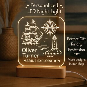 May include: A personalised LED night light with a nautical theme. The design features a sailboat, compass, and lighthouse, with the name "Oliver Turner" and "MARINE EXPLORATION" printed below. The light sits on a wooden base.