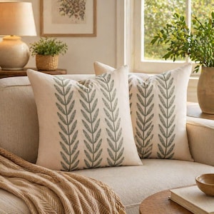 May include: Two square throw pillows with a cream-colored background and a repeating pattern of gray leaves. The pillows are on a beige sofa, with a tan throw blanket draped over the arm. A lamp and a small potted plant are visible in the background.