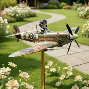Può includere: Un modello di aereo con un design mimetico, con una figura di pilota con occhiali. L'aereo ha un'elica ed è montato su un supporto dorato. Il testo "IRON MAIDEN" è visibile sul lato. Lo sfondo include fiori e un sentiero erboso.
