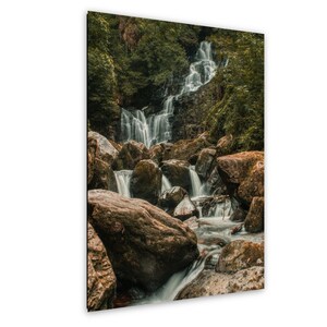 May include: A scenic photograph of a cascading waterfall flowing over rocks. The water is a bright white against the dark brown rocks and green foliage. The image is a vertical rectangle.