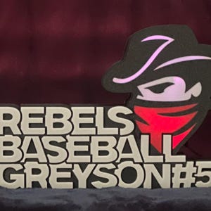 Op de afbeelding: Een honkbalbord met de woorden "REBELS BASEBALL GREYSON #5" in witte blokletters. Een zwart-rode afbeelding van een cowboy met een rode bandana en een zwarte hoed staat aan de rechterkant van het bord.