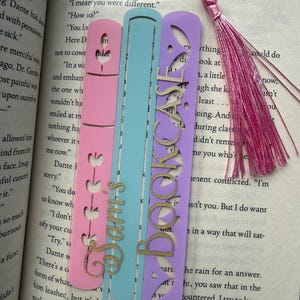 May include: A clear acrylic bookmark featuring three vertical cutouts in pink, light blue, and purple. The pink section has heart-shaped cutouts, the blue section is plain, and the purple section has the word BOOKCASE. A pink tassel is attached.