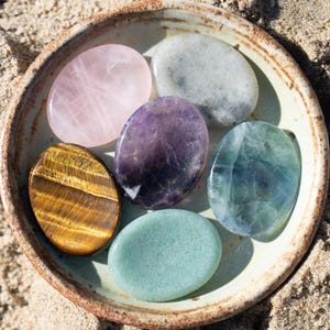 May include: A ceramic bowl filled with six polished gemstones of various colors, including pink, purple, green, and gold. The bowl sits on a sandy surface, with the gemstones arranged in a circular pattern. The gemstones are oval-shaped and appear smooth and reflective.