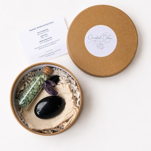 May include: A round brown cardboard gift box containing a glass vial filled with green aventurine chips, a polished black obsidian stone, a crescent-shaped amethyst, and a small beige fabric pouch, with an informational card and lid.