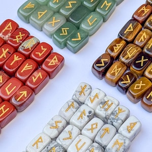 May include: A collection of polished rectangular stones arranged in four distinct groups. Each stone features a gold-colored engraved rune symbol. The sets include green, red, brown tiger's eye, and white marbled stones, all displaying unique natural patterns.
