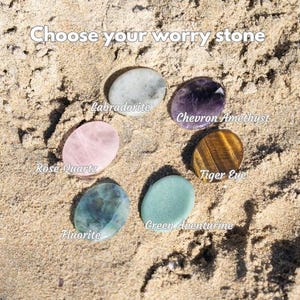 Puede incluir: Una selecci&oacute;n de piedras de preocupaci&oacute;n, incluyendo Labradorita, Amatista Chevron, Ojo de Tigre, Aventurina Verde, Fluorita y Cuarzo Rosa, dispuestas sobre arena. El texto "Choose your worry stone" es visible.