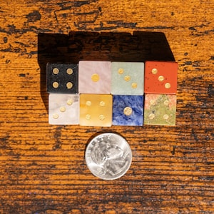 May include: A collection of seven square-shaped dice crafted from various stones, including black, pink, green, red, white, yellow, blue, and mottled green, each featuring gold-colored pips, positioned next to a silver United States quarter coin.